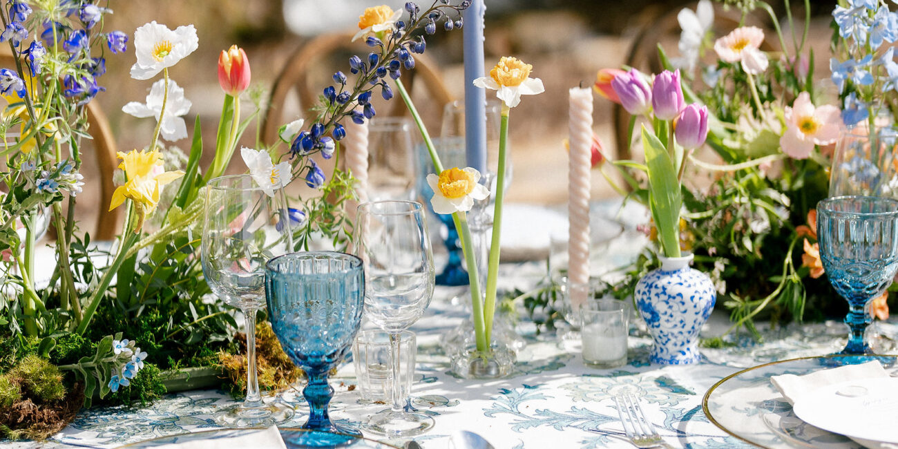 Table setting for an outdoor event, featuring a variety of flowers, glassware, and a patterned tablecloth